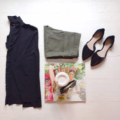 Image shows a folded pair of pants, black pointed toe shoes, a navy one-shoulder dress, and a stack of bridal magazines with sunglasses and a to-go cup of coffee on top.