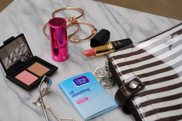 Image shows assorted makeup, face wipes, eyelash curler, jewelry, and a stylish black and white striped bag.